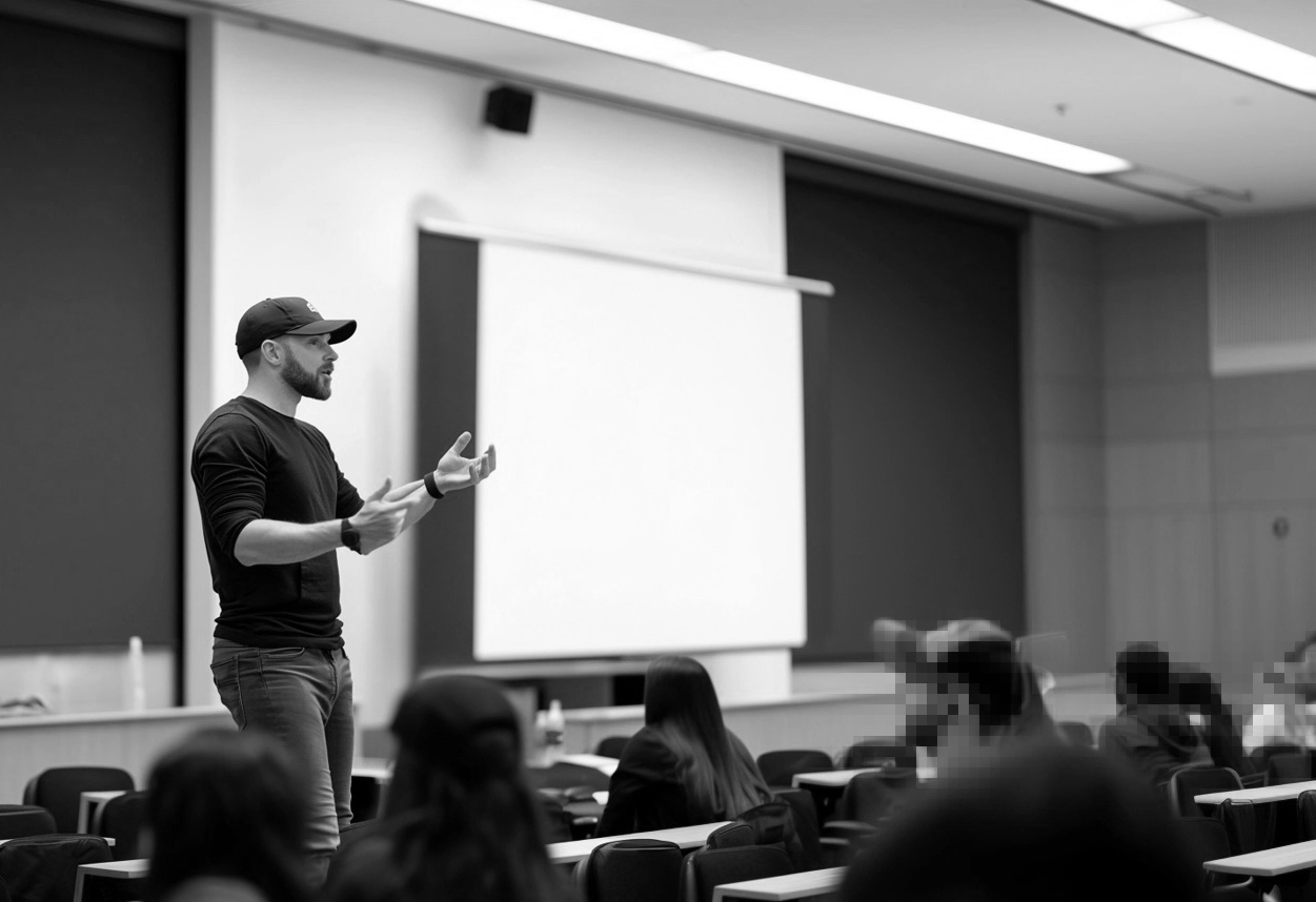 Man in casual clothes and cap speaking and gesturing in front of a classroom with seated students.