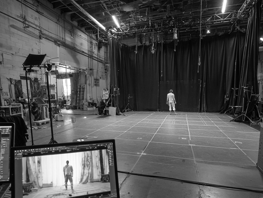 Person standing in an empty motion capture studio with grid-marked floor and black curtains, viewed from behind computer screens showing the person.